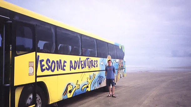 Man standing next to a yellow bus stopped on a beach.