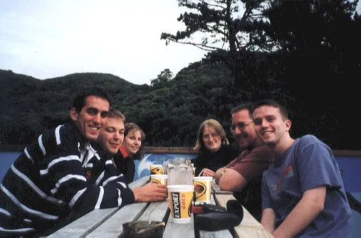 Six people sitting at a picnic table with beer cups and trees surrounding.