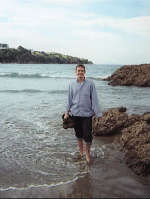 Man paddling on a beach holding his shoes with rocks close by.