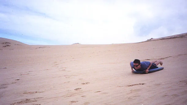 Man on his stomach riding a boogie board down a sand dune.